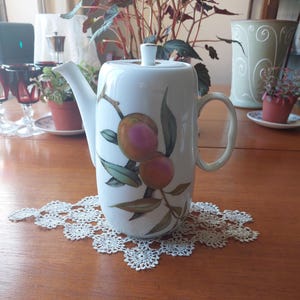 May include: White ceramic teapot with a peach and leaf design. The teapot has a lid and a handle. The design features two peaches with pink and orange hues, along with green leaves. The teapot is placed on a white lace doily.