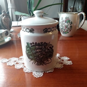 以下が含まれることがあります： 緑と茶色のハーブのフローラルデザインが施された蓋付きの白いセラミック製のキャニスター。キャニスターの上部にはロープのようなデザインが施されています。