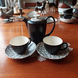 Könnte beinhalten: Ein schwarzes Keramik-Kaffeeservice mit einer Teekanne, zwei Tassen und Untertassen. Die Untertassen haben ein grau-weißes Muster. Die Tassen haben weiße Innenräume. Das Set steht auf einem Holztisch.