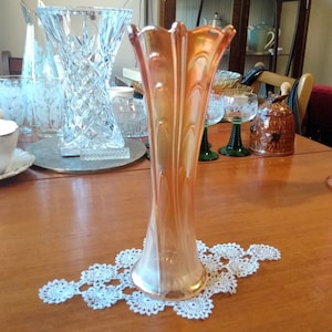 Puede incluir: Un jarrón de vidrio rosa iridiscente alto y acampanado, con un delicado encaje blanco debajo. El jarrón está sobre una mesa de madera.