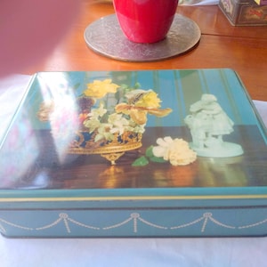Puede incluir: Una caja de lata rectangular vintage con fondo turquesa y diseño floral en la tapa. La tapa presenta un jarrón dorado adornado con flores y una pequeña figura blanca. La caja tiene un borde decorativo.