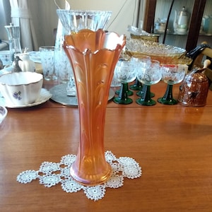 Puede incluir: Un jarrón de vidrio naranja iridiscente y alto, con un diseño acanalado. El jarrón está sentado sobre un encaje de ganchillo blanco en una mesa de madera.