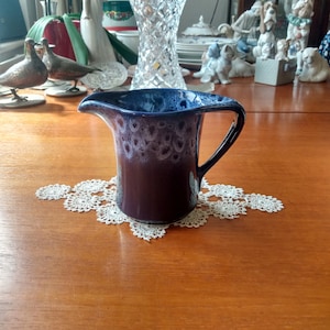 May include: A small, blue and brown ceramic creamer with a white glaze. The creamer has a handle and is sitting on a white doily.