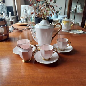 Puede incluir: Un juego de té vintage rosa y dorado, que incluye una tetera, una crema, una azucarera y tazas de té con platillos. El juego está dispuesto sobre una mesa de madera, mostrando su elegante diseño y atractivo clásico. La tetera tiene una altura aproximada de 25 cm.