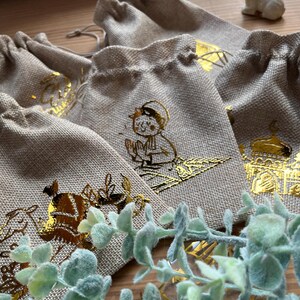 Könnte beinhalten: Fünf kleine, beigefarbene Jutesäckchen mit goldenen Foliendesigns. Die Designs beinhalten eine betende Person, eine Moschee und ein Kamel. Die Säcke liegen auf einer Holzoberfläche.