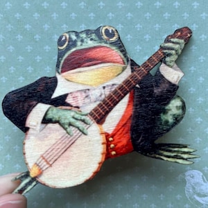 Könnte beinhalten: Ein grüner Frosch in einem Smoking und einer roten Weste spielt Banjo. Der Frosch hat einen weit geöffneten Mund und singt. Das Banjo ist weiß mit einem braunen Holzgriff.