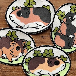 Op de afbeelding: Vier geborduurde patches met cartoon cavia's met klaverbladeren. De cavia's zijn bruin, zwart en wit met verschillende patronen.