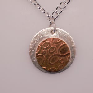May include: A silver necklace with a circular pendant. The pendant features a textured silver outer ring and a copper-colored inner circle with an organic pattern. The chain is a simple curb style.