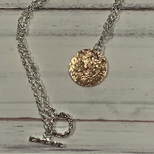 以下が含まれることがあります： ハンマーで叩いたゴールドの円形ペンダントが付いたシルバーチェーンネックレス。ネックレスはトグル留め具です。