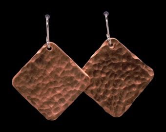 May include: A pair of copper earrings featuring a diamond-shaped design with a hammered, textured surface. Each earring is suspended from a simple silver-toned metal hook, creating a dangling effect with a warm, metallic finish and distinct indentations.