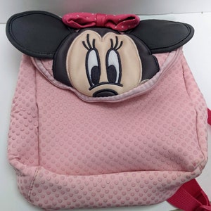 Op de afbeelding: Roze polka dot rugzak met een Minnie Mouse gezicht op de voorkant. De rugzak heeft een zwarte rits en een rode band.