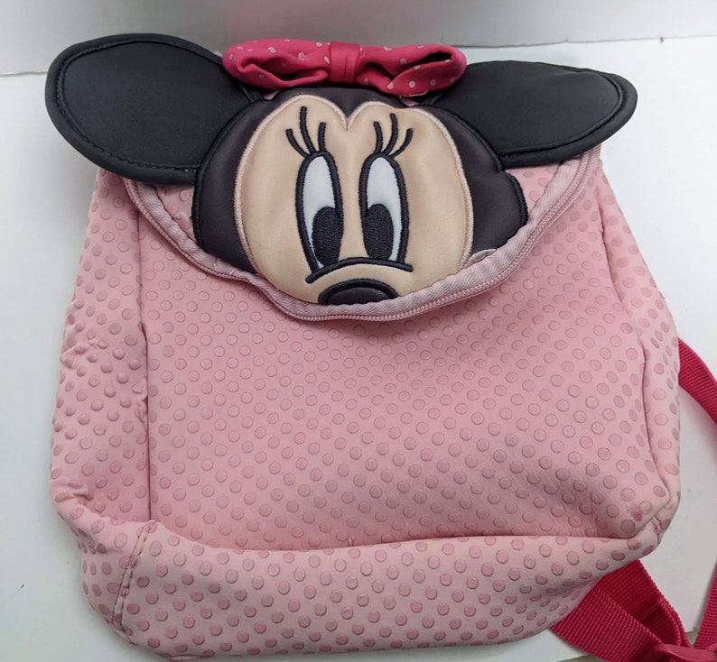 Op de afbeelding: Roze polka dot rugzak met een Minnie Mouse gezicht op de voorkant. De rugzak heeft een zwarte rits en een rode band.