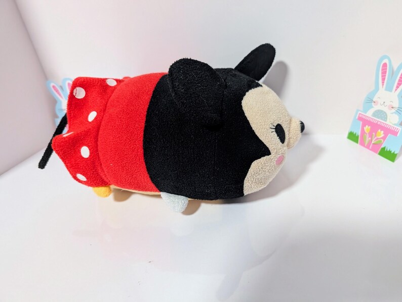 Peut inclure: Un jouet en peluche de Minnie Mouse portant une robe rouge &agrave; pois blancs. Le jouet est couch&eacute; sur le c&ocirc;t&eacute;, la t&ecirc;te tourn&eacute;e vers la droite. Le jouet a des oreilles noires et un nez noir.