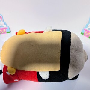 Peut inclure: Un jouet en peluche de Mickey Mouse dans sa tenue rouge et noire classique. Le jouet est couch&eacute; sur le dos, la t&ecirc;te tourn&eacute;e sur le c&ocirc;t&eacute;. Le jouet a un visage jaune et des yeux noirs.