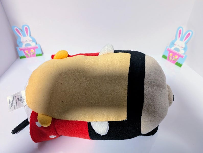 Peut inclure: Un jouet en peluche de Mickey Mouse dans sa tenue rouge et noire classique. Le jouet est couch&eacute; sur le dos, la t&ecirc;te tourn&eacute;e sur le c&ocirc;t&eacute;. Le jouet a un visage jaune et des yeux noirs.