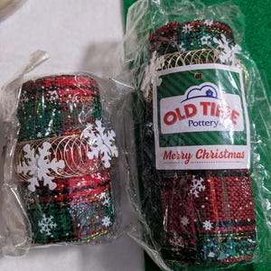 May include: Two rolls of red and green plaid ribbon with white snowflakes and gold wire. The ribbon is packaged in clear plastic bags. The larger roll has a label that reads "Old Time Pottery" and "Merry Christmas".