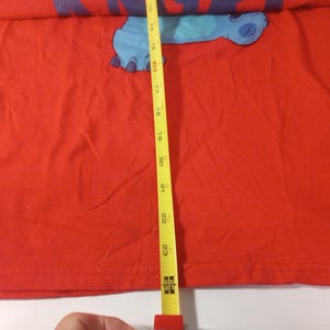 Puede incluir: Camiseta roja con un personaje de dibujos animados azul en la parte delantera. El personaje es un extraterrestre azul con una cabeza grande y ojos grandes. La camiseta se mide con una cinta m&eacute;trica amarilla, que muestra una longitud de 58 cm.