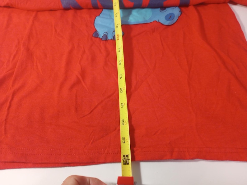 Puede incluir: Camiseta roja con un personaje de dibujos animados azul en la parte delantera. El personaje es un extraterrestre azul con una cabeza grande y ojos grandes. La camiseta se mide con una cinta m&eacute;trica amarilla, que muestra una longitud de 58 cm.