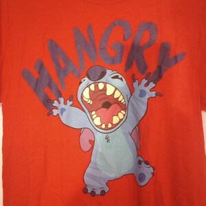 Puede incluir: Camiseta roja con un personaje azul de Stitch de Lilo & Stitch de Disney. Stitch est&aacute; gritando "HANGRY" en letras azules.
