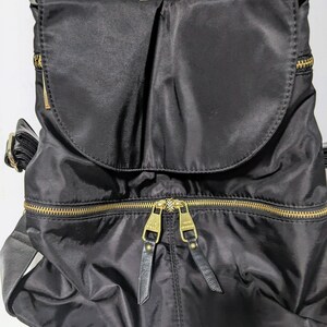 May include: Black nylon backpack with gold zippers and leather accents. The backpack has a flap closure and a top handle.