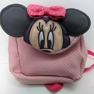 Op de afbeelding: Roze Minnie Mouse rugzak met zwarte oren en een roze stippen strik. De rugzak heeft een textuur met stippen.