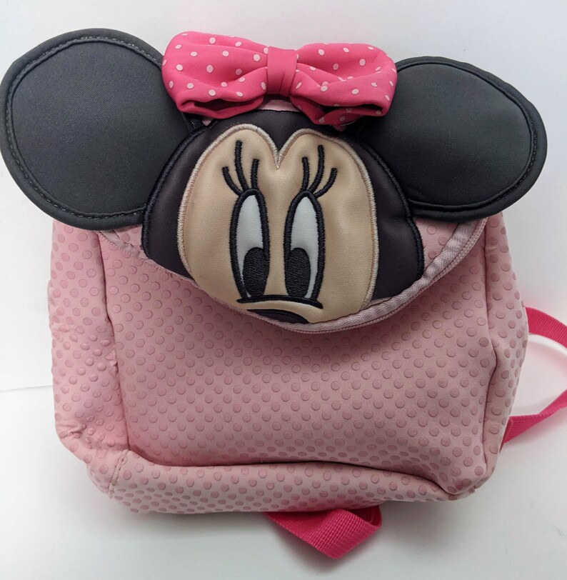 Op de afbeelding: Roze Minnie Mouse rugzak met zwarte oren en een roze stippen strik. De rugzak heeft een textuur met stippen.