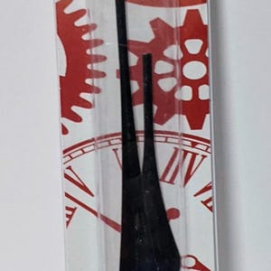 May include: Black clock hands packaged in a clear plastic sleeve with a red and white background featuring clock faces and gears.