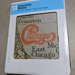 May include: A white 8-track tape cartridge with a blue and white Columbia House label. The front of the cartridge features a map of Chicago with the band name "Chicago" in a large, stylized font.