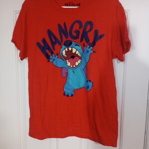 Puede incluir: Camiseta roja con un personaje azul Stitch de Lilo & Stitch. La camiseta tiene la palabra "HANGRY" impresa en letras negras.