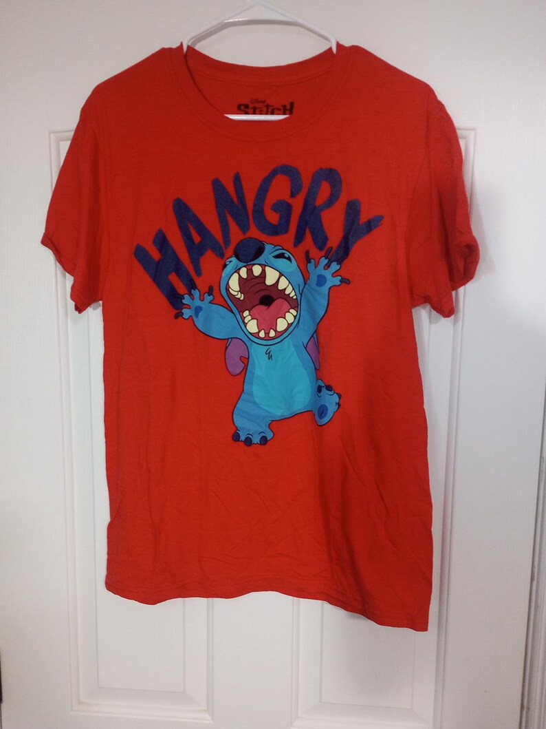 Puede incluir: Camiseta roja con un personaje azul Stitch de Lilo & Stitch. La camiseta tiene la palabra "HANGRY" impresa en letras negras.