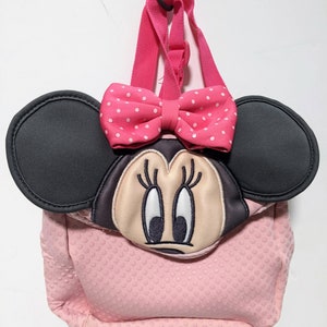 Op de afbeelding: Roze Minnie Mouse rugzak met zwarte oren en een roze strik met witte stippen. De rugzak heeft een roze textuur met kleine witte stippen.