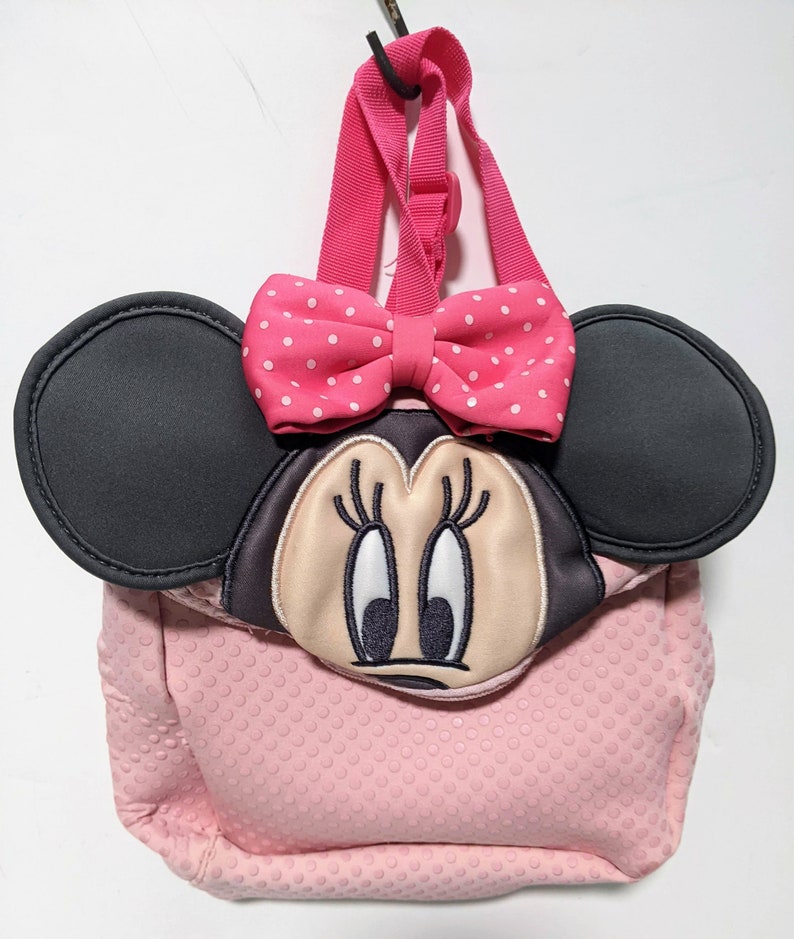 Op de afbeelding: Roze Minnie Mouse rugzak met zwarte oren en een roze strik met witte stippen. De rugzak heeft een roze textuur met kleine witte stippen.