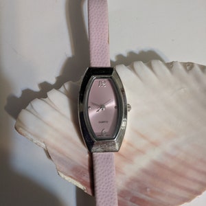 May include: A pink leather wristwatch with a silver oval-shaped face and a quartz movement. The watch is on a white seashell.