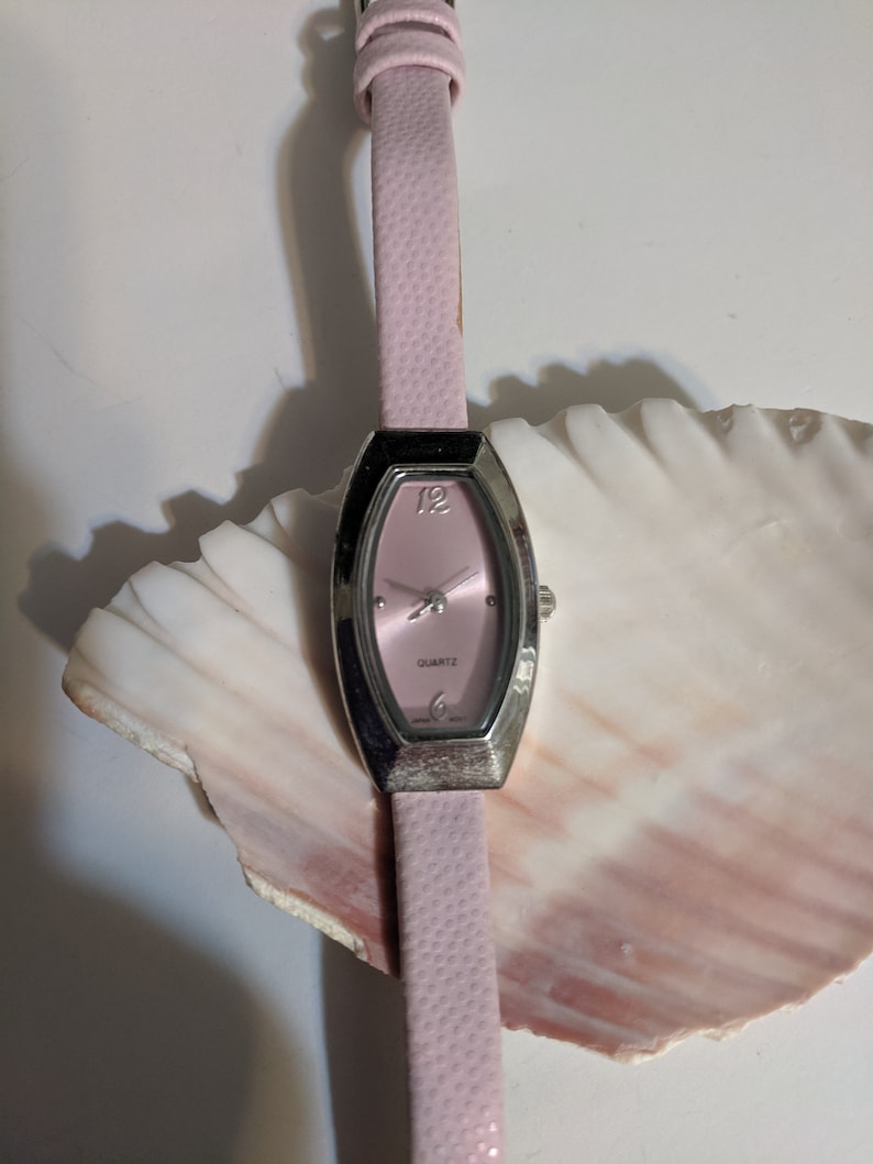 May include: A pink leather wristwatch with a silver oval-shaped face and a quartz movement. The watch is on a white seashell.