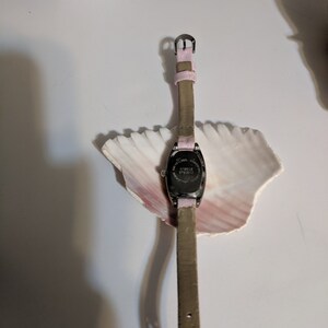 May include: A silver-toned watch with a pink leather band and a rectangular face. The watch is on a white surface with a seashell.