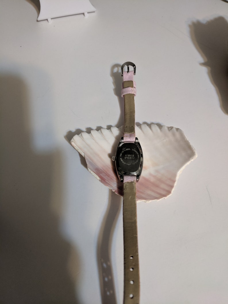 May include: A silver-toned watch with a pink leather band and a rectangular face. The watch is on a white surface with a seashell.
