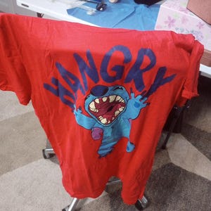 Puede incluir: Camiseta roja con un personaje de dibujos animados azul con la palabra "HANGRY" en letras azules.