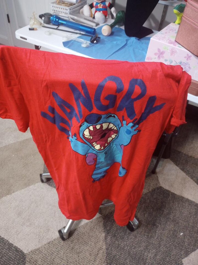 Puede incluir: Camiseta roja con un personaje de dibujos animados azul con la palabra "HANGRY" en letras azules.
