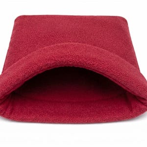May include: A deep red, plush, fleece-like pet sleeping pouch with a wide, rounded opening. The exterior and interior feature a soft, textured fabric surface. The structure is semi-rigid, maintaining a curved, tunnel-like shape for resting.