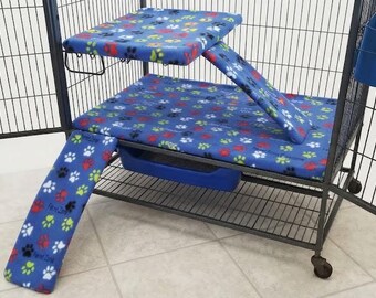 May include: A black wire pet cage featuring a blue fabric liner with a colorful paw print pattern. The cage includes a raised platform, a ramp leading to the platform, a second ramp, and a blue plastic tray and hanging container attached to the side.