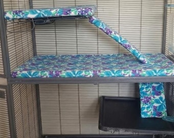 May include: A set of pet cage accessories featuring a floral pattern in shades of purple, teal, and white. The set includes a rectangular platform cover, a matching ramp cover, a large base mat, and a smaller rectangular mat for a lower level.