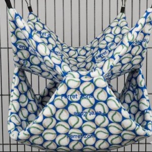 May include: A double-decker fabric hammock for small animals featuring a white and blue baseball pattern with green stitching details. The soft fleece material is printed with the text Ferret Zone throughout and hangs from a metal wire cage.