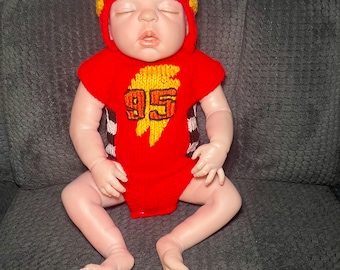 Puede incluir: Un conjunto de bebé de ganchillo que consta de un gorro rojo con capucha con detalles de ojos blancos y azules y un body rojo a juego con un gráfico de rayo amarillo con el número 95 en negro y naranja, usado por una muñeca realista.