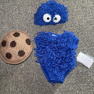 May include: A three-piece baby outfit set. The set includes a blue, fuzzy hat with large white eyes, a blue fuzzy diaper cover, and a brown crocheted hat with chocolate chip details. The outfit is designed to resemble a popular cartoon character.