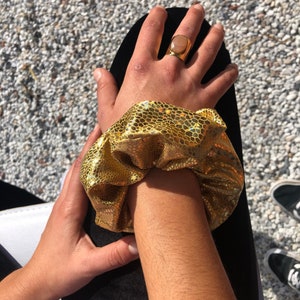 May include: A gold scrunchie with a textured, scale-like pattern is wrapped around a wrist. A gold ring with a light pink stone is on a hand. The background is a gravel surface.