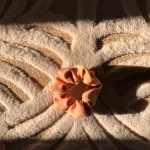May include: A peach-colored scrunchie made of soft fabric. The scrunchie is lying on a white and brown patterned rug.
