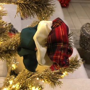 May include: Three scrunchies in a Christmas tree. One is dark green, one is cream-colored, and the third is red and black plaid. The tree is gold and glittery, with a white background.