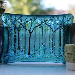 May include: A blue glass dish featuring a forest scene. The dish is decorated with small blue, green and white glass pieces. The forest scene is made up of black trees and a white background.