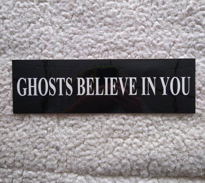Rectangular sticker with black background and white lettering. The text reads ghosts believe in you.
