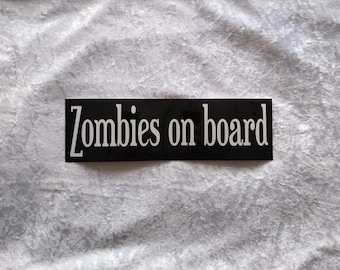 Op de afbeelding: Zwart-witte sticker met de tekst "Zombies on board" in wit gedrukt.